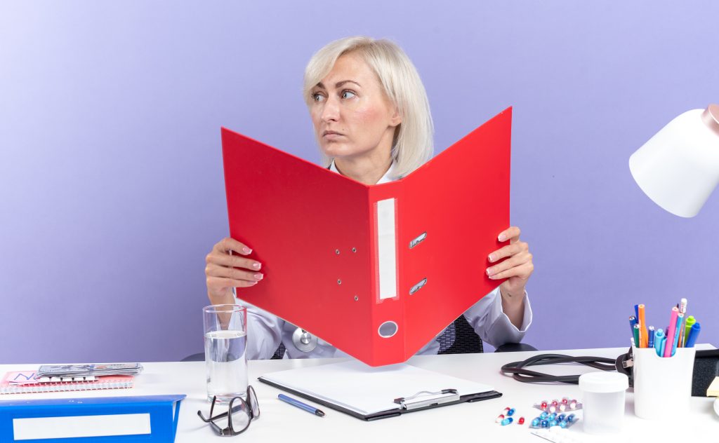 The Simple Way to Stay Compliant in 2026 Without Losing Your Mind 6 confident adult slavic female doctor medical robe with stethoscope sitting desk with office tools holding file folder looking side isolated purple background with copy space